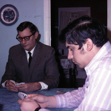 Two men sitting at a table, one playing cards, the other observing intently.
