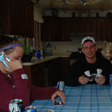Two people sitting at a table in a kitchen, one looking down, the other holding glasses.