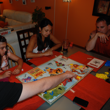 Four people playing a board game at a table, with colorful game pieces and snacks nearby.