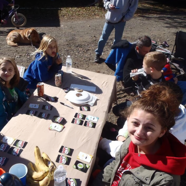 Group of children and adults gathering around a table, enjoying snacks in an outdoor setting.