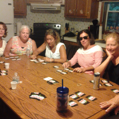 A group of six women gathered around a table, playing cards and socializing in a kitchen.