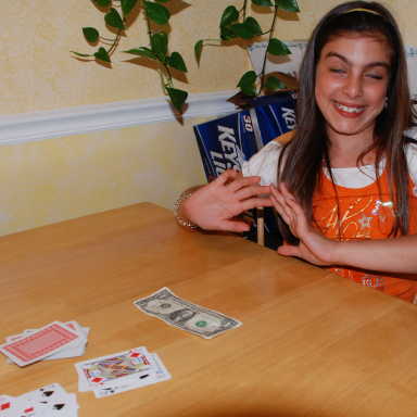 A girl smiles while playing cards at a table, with money and cards in front of her.