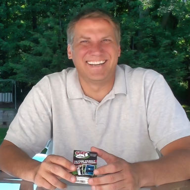 Smiling man holding a pack of batteries outdoors, surrounded by greenery.