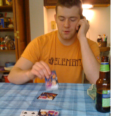 A young man on the phone plays cards at a table with a drink nearby.