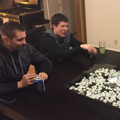 Two men sitting at a table playing a game with white pieces and drinks nearby.